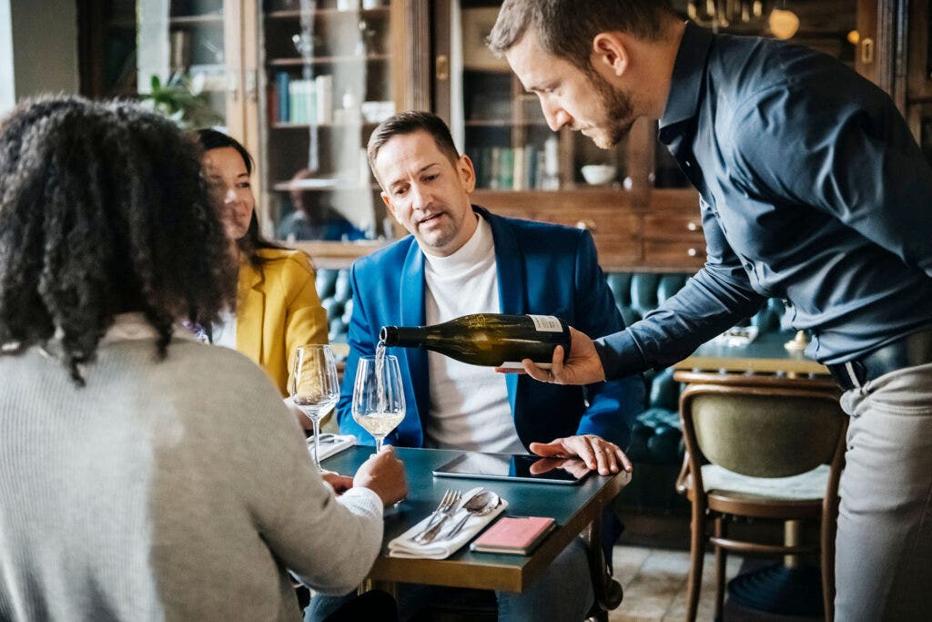 A waiter delicately pouring some white wine for a group of customers in a fancy restaurant.